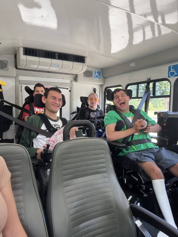Four students seated in wheelchairs ride together on an accessible bus, smiling and laughing during an outing.