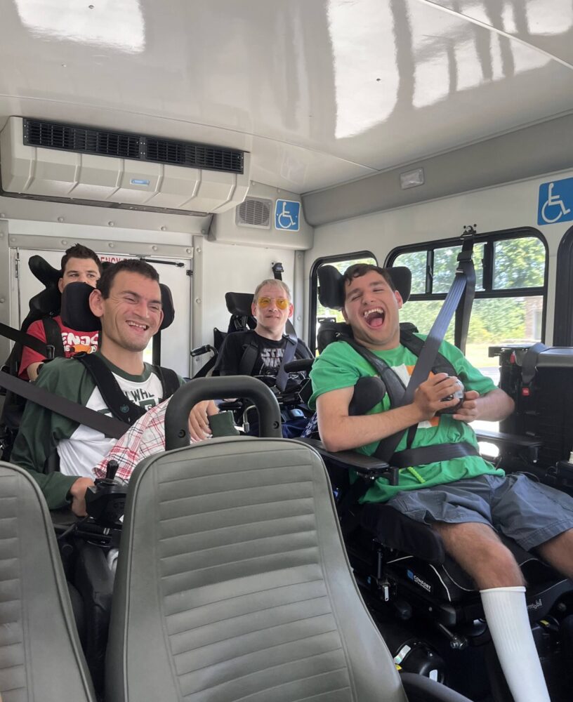 Four students seated in wheelchairs ride together on an accessible bus, smiling and laughing during an outing.
