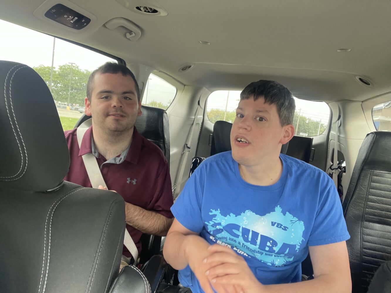 Two residents sit side by side in the back of a van, both wearing seatbelts; one wears a maroon polo shirt and looks calmly toward the camera, while the other wears a blue T-shirt and gestures with their hands as they look ahead, with daylight visible through the vehicle windows.