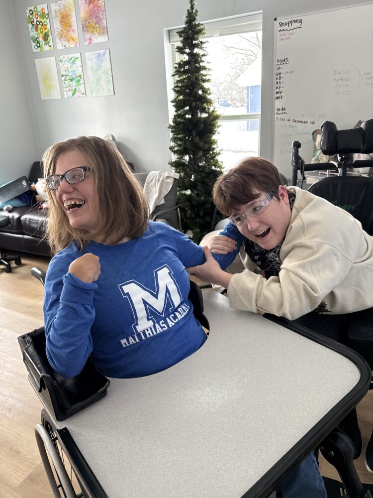 Two students seated at a table smiling and laughing together during a classroom activity, leaning toward each other in a bright room.
