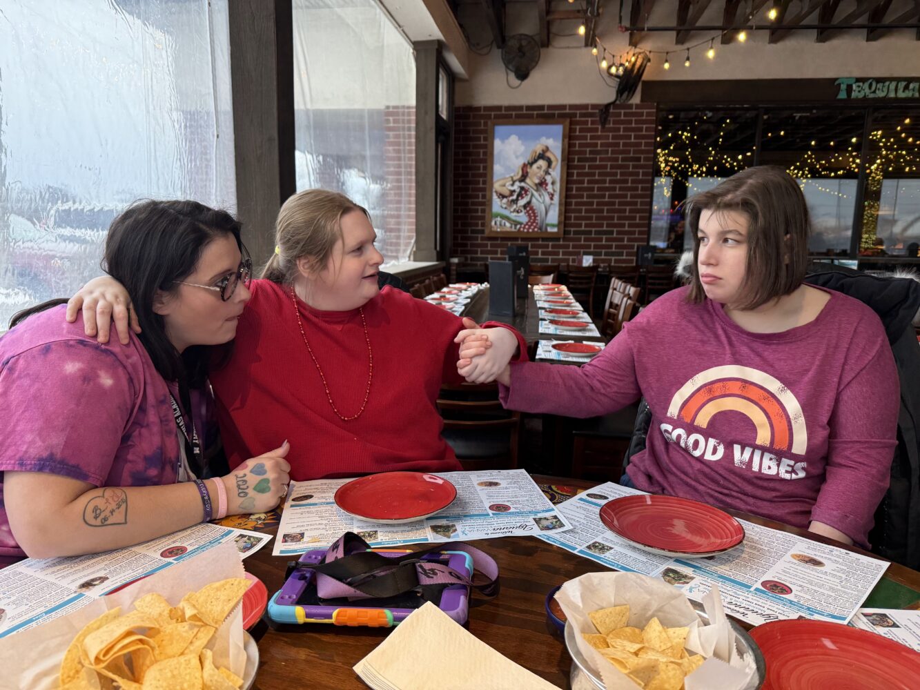 Three women sit together at a restaurant table with menus and red plates; two hold hands across the table while the third leans in close, creating a warm, supportive moment as baskets of tortilla chips sit in front of them under string lights.